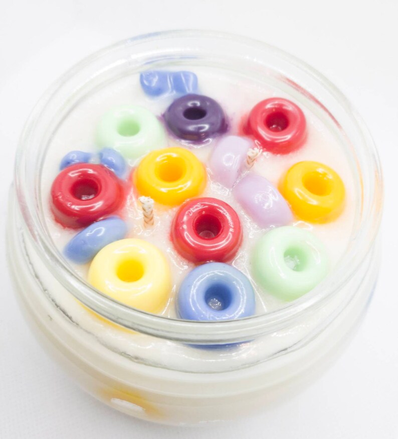 Pu&ograve; includere: Una candela in barattolo di vetro con inserti di cera colorati a forma di anello nei toni del rosso, giallo, verde, blu e viola. La candela ha due stoppini e una base di cera bianca cremosa. Sfondo bianco.