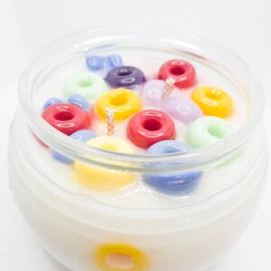 Pu&ograve; includere: Una candela in barattolo di vetro trasparente riempita di cera bianca e pezzi di cera colorati a forma di anello. Gli anelli sono di vari colori, tra cui rosso, giallo, verde, viola e blu. Sono visibili due stoppini.