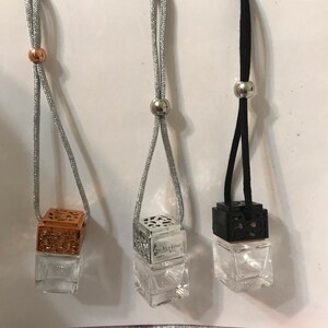 May include: Three hanging car air fresheners. One has a copper-colored top, one has a silver top, and one has a black top. Each has a clear glass cube-shaped bottle. The silver and black tops have a decorative openwork design. The fresheners hang from a silver or black cord.