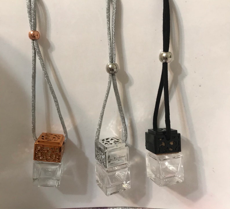 May include: Three hanging car air fresheners. One has a copper-colored top, one has a silver top, and one has a black top. Each has a clear glass cube-shaped bottle. The silver and black tops have a decorative openwork design. The fresheners hang from a silver or black cord.