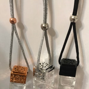 May include: Three car air fresheners with clear glass cube bottles. The tops are metallic in copper, silver, and black. Each is suspended by a cord with a metallic bead. The copper and silver tops have decorative cut-out patterns.