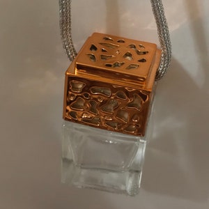 May include: A clear, square glass bottle with a decorative copper-colored top and a silver chain. The top has a patterned design. The bottle is empty and suspended by the chain.