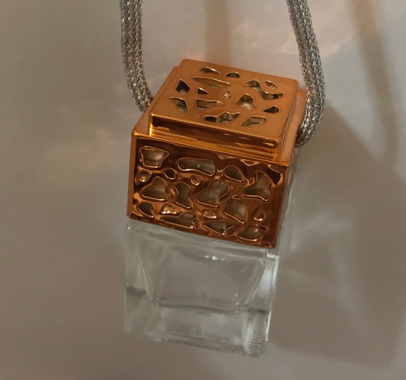 May include: A clear, square glass bottle with a decorative copper-colored top and a silver chain. The top has a patterned design. The bottle is empty and suspended by the chain.