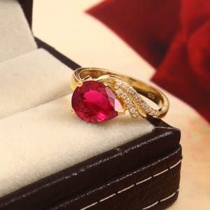 Op de afbeelding: Een gouden ring met een grote, peervormige, helderrode edelsteen. De ring heeft een band versierd met kleine, heldere accentstenen. De ring wordt gepresenteerd in een zwarte sieradendoos met een witte binnenkant.