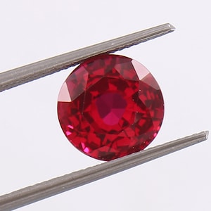 Puede incluir: Una piedra preciosa de color rojo intenso con forma redonda y superficie facetada. La piedra preciosa está sujeta por unas pinzas.