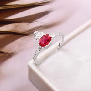 May include: A silver ring featuring a large oval-cut ruby gemstone. The ring has a small cluster of three round stones above the ruby and smaller stones set along the band. The ruby is a vibrant red colour.