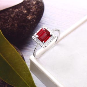 Könnte beinhalten: Ein silberner Ring mit einem quadratischen, tiefroten Edelstein. Der Edelstein ist in einem quadratischen Rahmen gefasst, der von kleineren, klaren Steinen umgeben ist. Das Ringband hat ein gedrehtes Seildesign, das Textur verleiht.