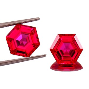 May include: Two faceted, hexagonal, bright red gemstones. One is held by tweezers, the other rests on a reflective surface. The gems have a complex internal structure, with multiple facets reflecting light.