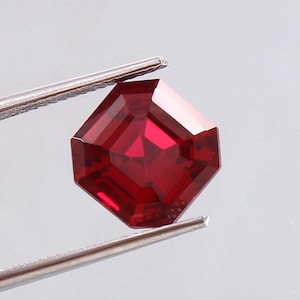 May include: A square-cut, deep red gemstone is held by silver tweezers. The gemstone has an octagonal shape with multiple facets that reflect light. The gem is a vibrant, saturated red colour. The tweezers are made of metal.