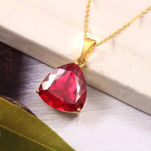Pode incluir: Um colar dourado com um pingente triangular facetado de pedra preciosa vermelha escura. O pingente está suspenso por uma delicada corrente dourada. A pedra preciosa é de uma cor vermelha vibrante e chamativa.
