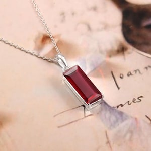 Puede incluir: Un collar de plata con un colgante rectangular de piedra preciosa rojo oscuro. El colgante está engastado en un marco de plata y cuelga de una delicada cadena de plata. La piedra preciosa tiene una superficie pulida y el collar se muestra sobre un fondo suave y borroso.