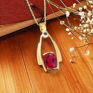 May include: A gold pendant necklace featuring a cushion-cut deep red gemstone set within a polished gold frame. A small, clear, round-cut stone is positioned at the top of the pendant, connecting to a delicate gold chain that hangs vertically.
