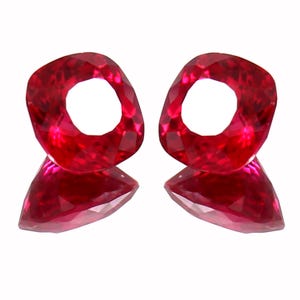 May include: Two faceted, heart-shaped, ruby-red gemstones with a hole in the center. The gems are displayed on a white background, showcasing their brilliant cut and color. The gems are likely for jewelry.