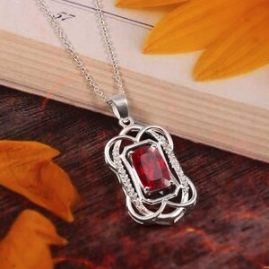 Pode incluir: Um colar de prata com um pingente de pedra vermelha. O pingente é rodeado por pedras transparentes menores e tem um design delicado e intrincado.