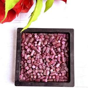 May include: A square black tray filled with a collection of small, raw, unpolished ruby crystals in various shades of pink and deep red. The stones are irregular in shape and size, creating a dense, textured layer within the container.