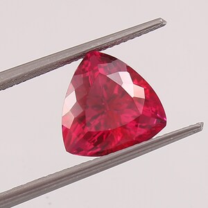 May include: A deep red triangular gemstone with a faceted cut. The gemstone is held in place by a pair of tweezers.