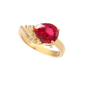 May include: A gold ring with a large, pear-cut, bright red gemstone. The ring's band is gold, with a section embellished with small, clear gemstones. The design is elegant and striking, suitable for special occasions.