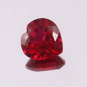 Può includere: Una gemma di rubino sfaccettata a forma di cuore. La gemma rosso intenso è altamente riflettente, con molteplici sfaccettature che catturano la luce. La gemma è esposta su una superficie riflettente, creando un effetto specchio.