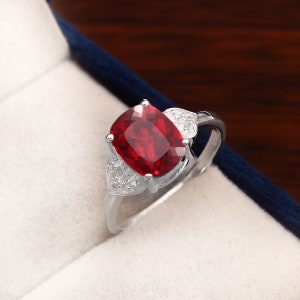 Puede incluir: Un anillo de plata con una gran piedra roja de talla cojín, rodeada de piedras más pequeñas y transparentes. El anillo está engastado en un delicado diseño en forma de hoja.
