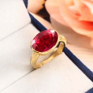 Op de afbeelding: Een gouden ring met een grote, ovale, rode edelsteen. De ring ligt in een blauwe en witte doos.