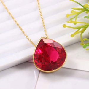 May include: A gold-colored necklace featuring a large, faceted, teardrop-shaped red gemstone pendant. The pendant is set in a gold bezel and hangs from a delicate gold chain. The background is white with a hint of green.