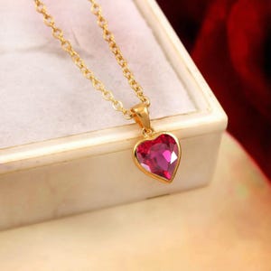 Könnte beinhalten: Eine goldfarbene Halskette mit herzförmigem Anhänger. Der Anhänger hat einen leuchtenden, rubinroten Edelstein in einer goldenen Lünette. Die Halskette wird auf einer weißen Oberfläche mit verschwommenem rotem Hintergrund präsentiert.