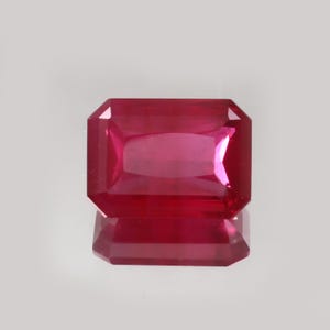 Puede incluir: Una gema de rubí de corte rectangular con un tono rojo intenso. La gema está facetada, con una superficie pulida que refleja la luz. El rubí se encuentra sobre un fondo neutro, resaltando su color y claridad. La reflexión de la gema es visible debajo.