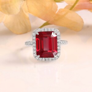 Puede incluir: Un anillo de plata con una piedra preciosa roja de talla esmeralda y un halo de pequeñas piedras blancas.