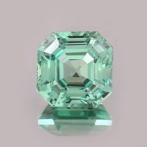 Könnte beinhalten: Ein facettierter, quadratisch geschliffener Edelstein in hellem Grün. Der Stein hat ein symmetrisches, mehrfach facettiertes Design, das Licht reflektiert. Der Edelstein wird auf einer reflektierenden Oberfläche präsentiert. Thema: Schmuck.