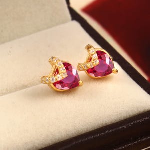 Peut inclure: Une paire de boucles d'oreilles à clous en or avec une pierre précieuse rouge en forme de cœur. Chaque boucle est ornée d'une bande incurvée de petites pierres blanches transparentes qui entourent le côté du cœur rouge pour un rendu fini.