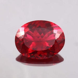 May include: A vibrant red oval gemstone with a faceted cut. The gemstone is set against a white background.