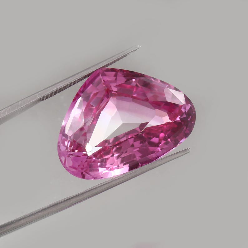 May include: A faceted, triangular pink gemstone is held by silver tweezers against a gray background. The gem has a high degree of clarity, with multiple facets reflecting light. The color is a vibrant, saturated pink.