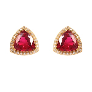 May include: A pair of stud earrings featuring triangular-cut deep red gemstones set in gold. Each central stone is surrounded by a halo of small, clear, round-cut white diamonds, creating a bright contrast against the vibrant red center.