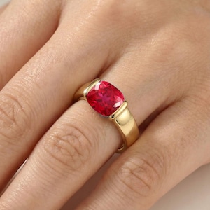 May include: A gold ring with a large, cushion-cut, bright red gemstone. The ring has a simple, elegant design with a polished gold band and bezel setting. The gemstone is the focal point, showcasing its rich colour and brilliance.
