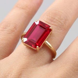 May include: A gold ring featuring a large, rectangular, emerald-cut red gemstone held in place by four prongs. The band is thin and polished gold, showcasing the vibrant, deep red color of the faceted stone which sits prominently on the finger.