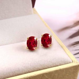 Könnte beinhalten: Ein Paar rubinrote, oval geschliffene Ohrstecker mit Edelsteinen, gefasst in Gold. Die Ohrringe werden in einer cremefarbenen Schmuckschatulle mit goldfarbenem Rand präsentiert. Die Edelsteine haben eine leuchtende, tiefrote Farbe.