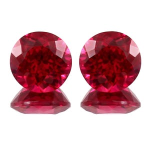 Puede incluir: Dos gemas redondas, facetadas, de color rojo intenso. Cada gema está colocada sobre una base facetada a juego, creando un efecto de espejo. Las gemas tienen un corte brillante, que refleja la luz. La presentación general es limpia y elegante, destacando el color y la claridad de las gemas.