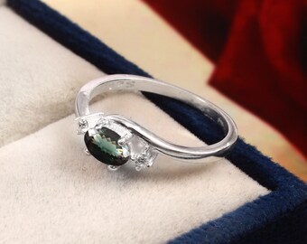 May include: A silver ring with a green oval gemstone and smaller clear gemstones. The ring is set in a white box.