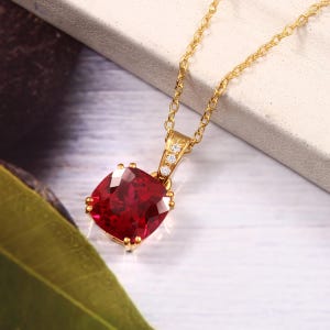 May include: A gold necklace with a square, deep red gemstone pendant. The pendant is adorned with small, clear stones. The chain is delicate, creating an elegant and striking design.