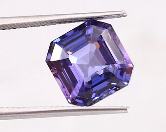 May include: A purple gemstone with a square emerald cut. The gemstone is held by tweezers against a white background.