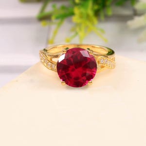 Könnte beinhalten: Ein goldfarbener Ring mit einem großen, runden, tiefroten Edelstein als Mittelpunkt. Das Band hat ein dekoratives Design mit kleinen, klaren Akzentsteinen. Der Ring ist vor einem neutralen Hintergrund platziert, der die lebendige Farbe des Edelsteins hervorhebt.