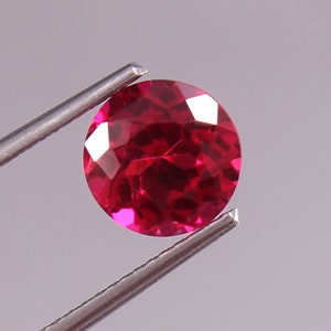 May include: A deep red gemstone with a round cut and many facets. The gemstone is held by a pair of tweezers.