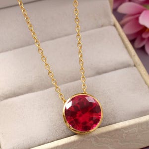 May include: A gold necklace with a large, round, faceted ruby pendant. The ruby is a bright red colour and set in a gold bezel. The chain is delicate and gold-coloured.