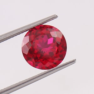 May include: A round, red gemstone with a faceted cut. The gemstone is held in place by a pair of tweezers.