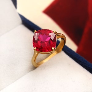 May include: A gold ring with a large, square-cut, bright red gemstone. The ring's band and setting are gold, contrasting with the deep red of the stone. The ring is displayed in a blue and white jewelry box.