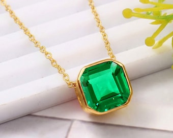 May include: A gold-toned necklace featuring a large, square-cut emerald green gemstone pendant. The pendant is set in a gold bezel and hangs from a delicate gold chain. The gemstone has a faceted surface, reflecting light.