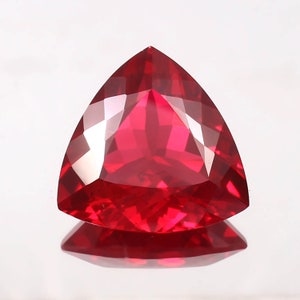 Puede incluir: Una gema facetada de forma triangular con un tono rojo intenso. La superficie de la gema refleja la luz, mostrando sus intrincados cortes y ángulos. La gema se exhibe sobre una superficie reflectante, realzando su brillo.