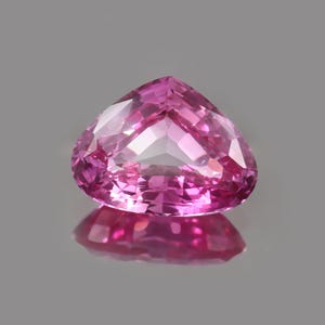 May include: A faceted, heart-shaped pink gemstone with a high degree of clarity. The gem reflects light, creating a brilliant display of color and sparkle. The gemstone sits on a reflective surface.