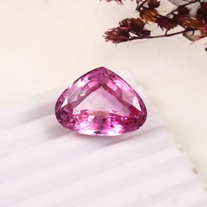 May include: A faceted, heart-shaped pink gemstone. The gem has a brilliant cut, reflecting light. The stone is set against a white background, with a hint of dried flowers in the upper right corner. The gem is a vibrant shade of pink.