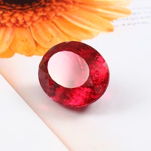 Puede incluir: Una piedra preciosa facetada de forma ovalada con un color rojo intenso y vibrante. La piedra presenta una superficie pulida con múltiples cortes geométricos que reflejan la luz, creando una apariencia brillante. La piedra es translúcida y carmesí.
